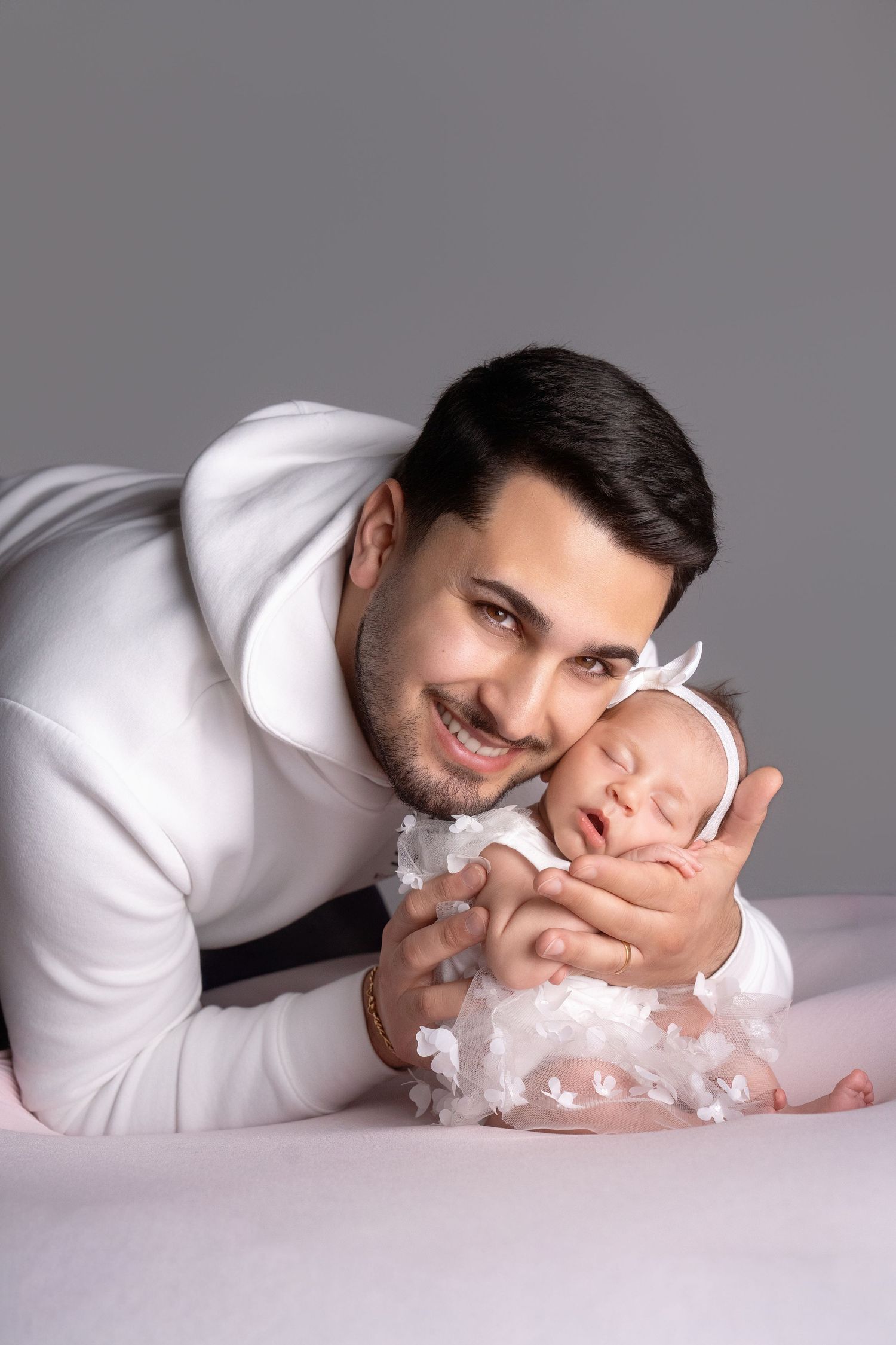Newborn baby in white lace outfit lying on grey studio backdrop.