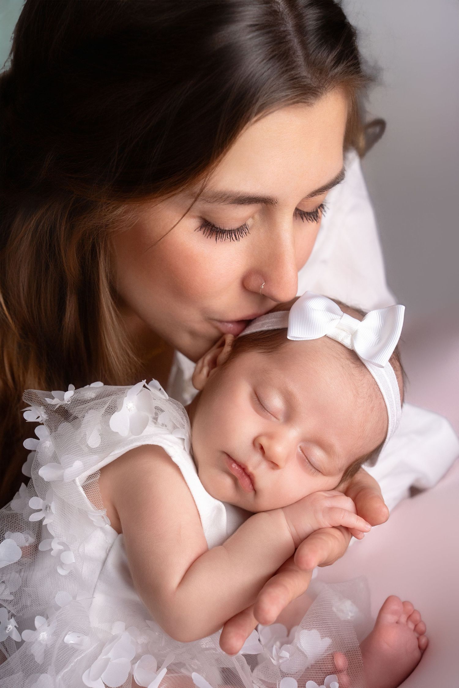 Mother tenderly kissing sleeping baby in white lace dress.