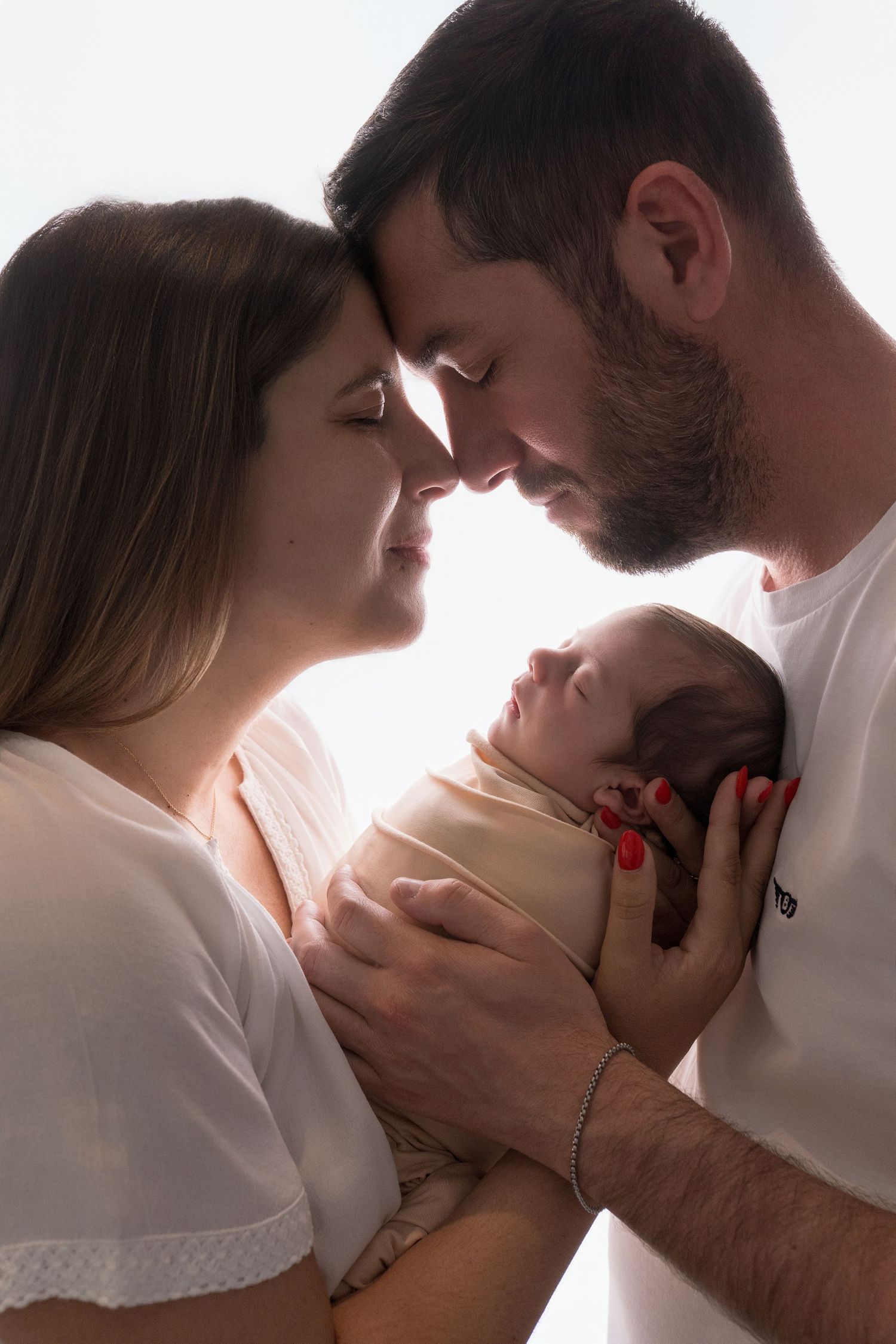 New parents tenderly hold their swaddled newborn baby together, touching foreheads in an intimate family portrait.