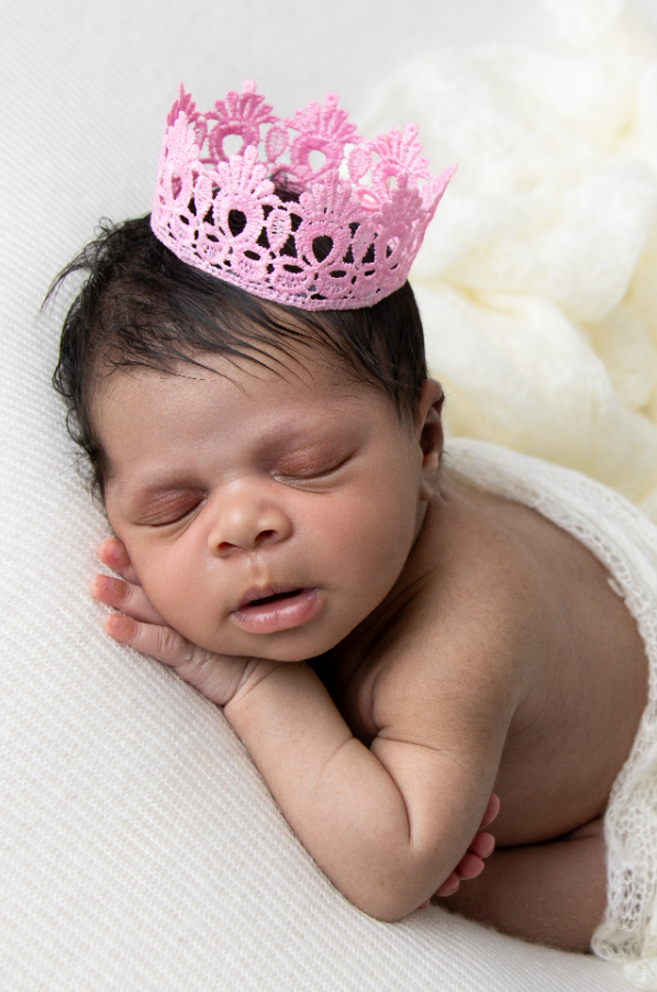 Sleeping newborn wearing a delicate pink crown rests on white fabric.