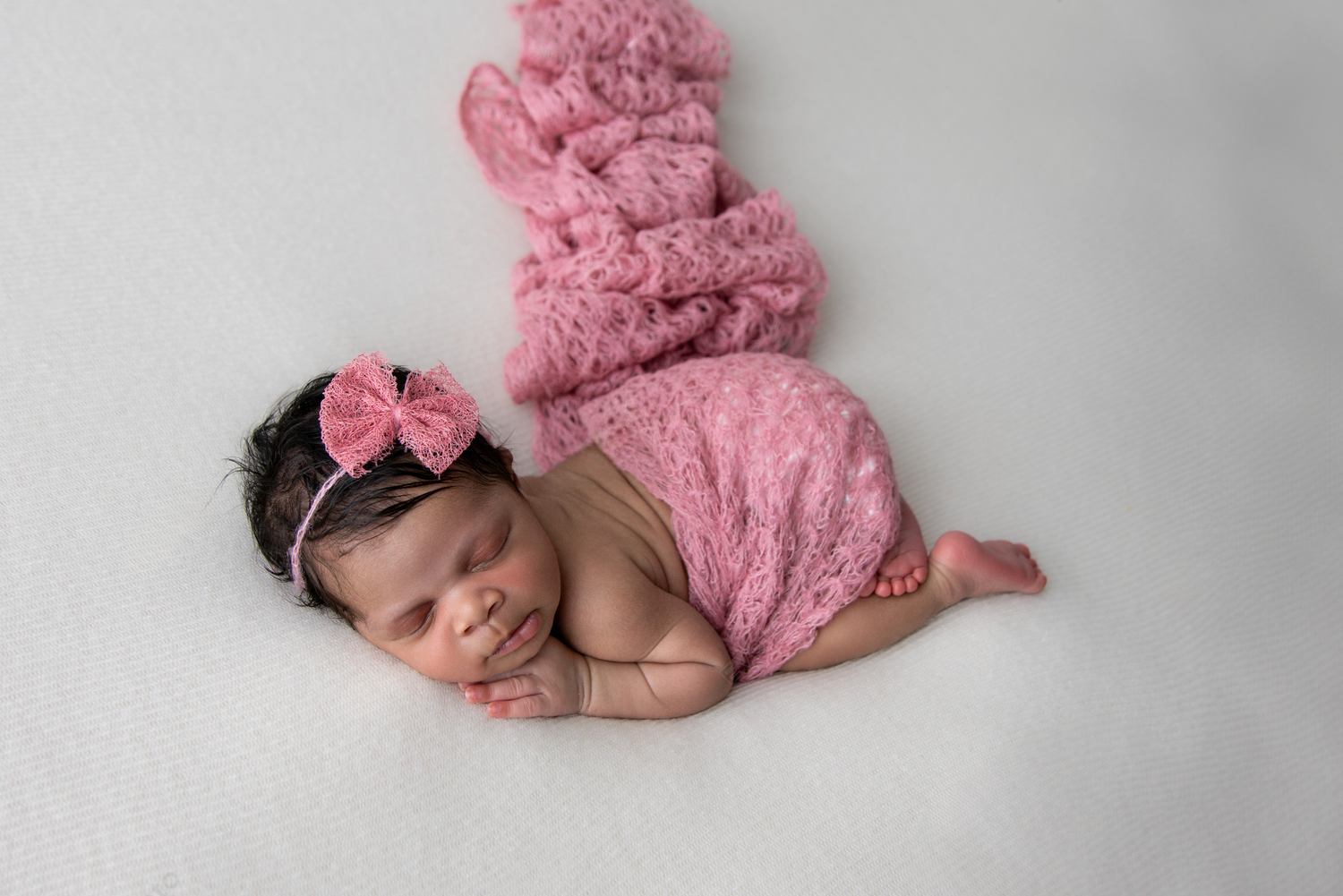 Newborn baby wearing pink fuzzy accessories sleeps peacefully on grey background.