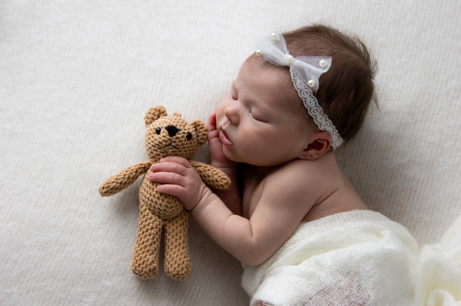 Sleeping baby with white headband poses with small teddy bear on white backdrop.