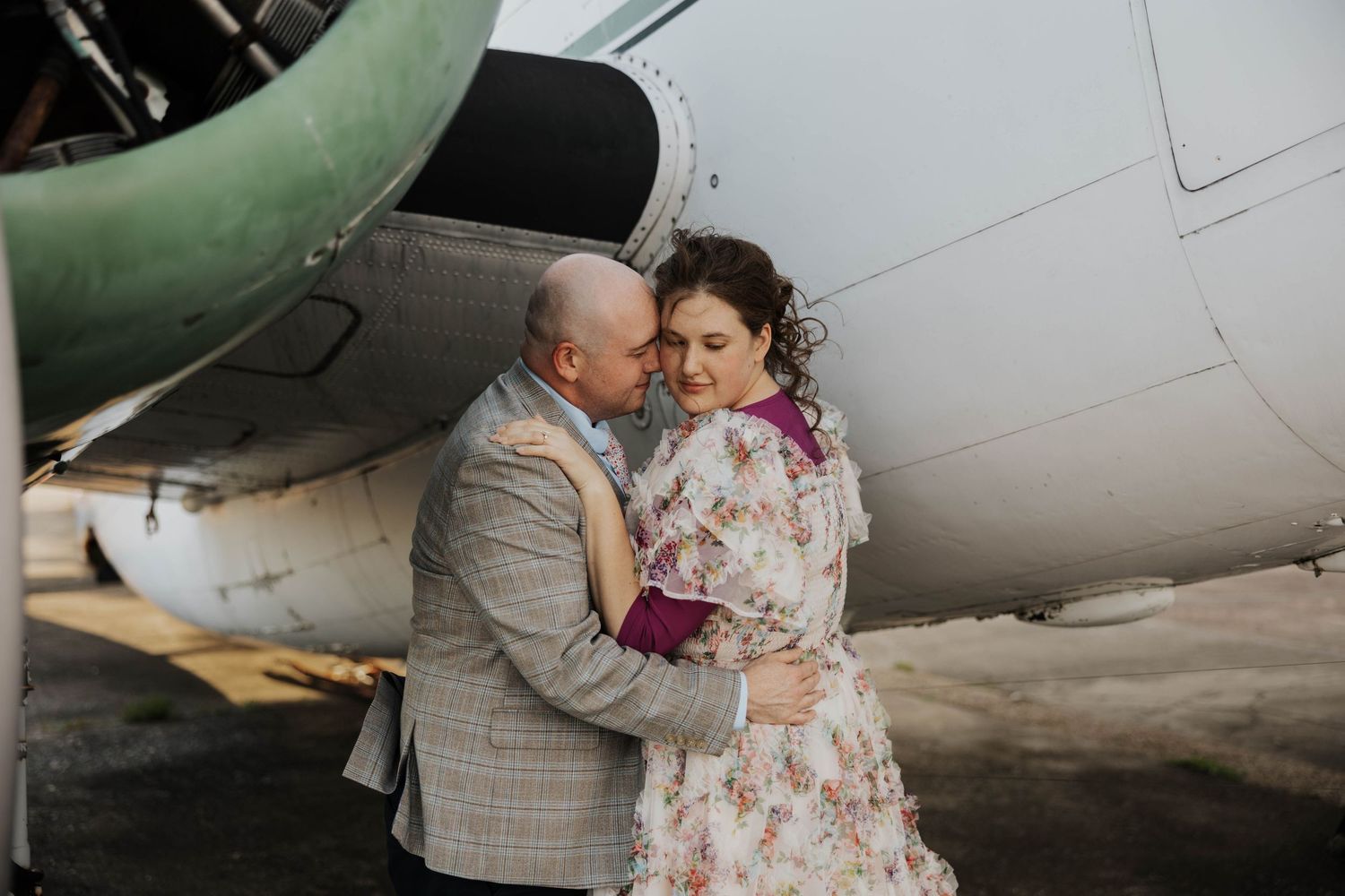 A romantic couple embraces next to a vintage aircraft with the groom wearing a gray suit and bride in floral dress.