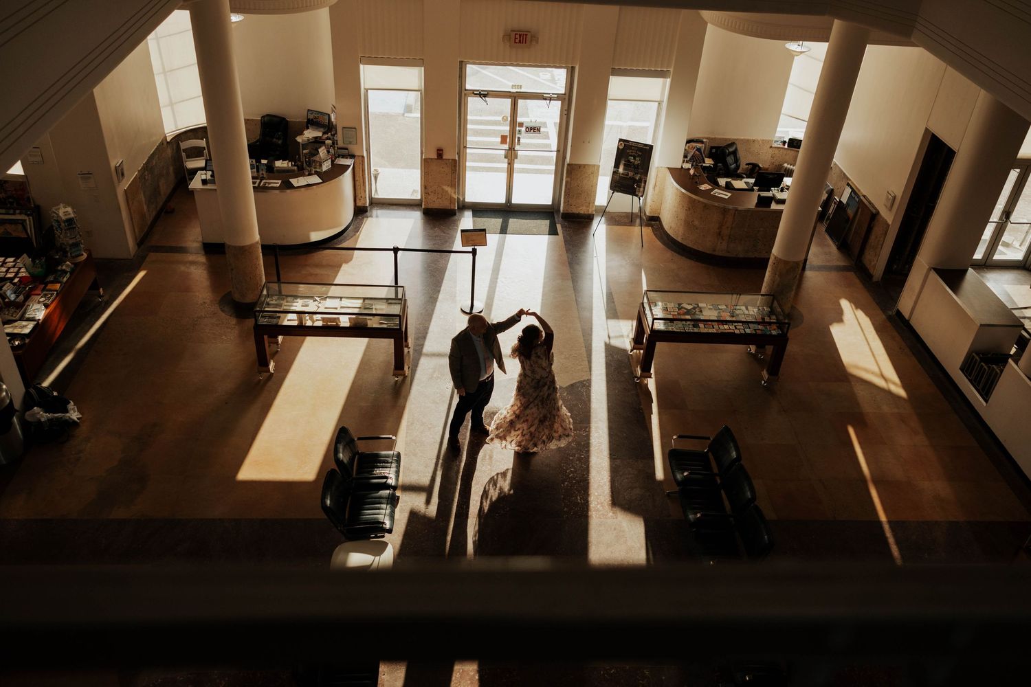 A silhouetted couple shares an intimate dance in a grand hall with natural light streaming through tall windows.