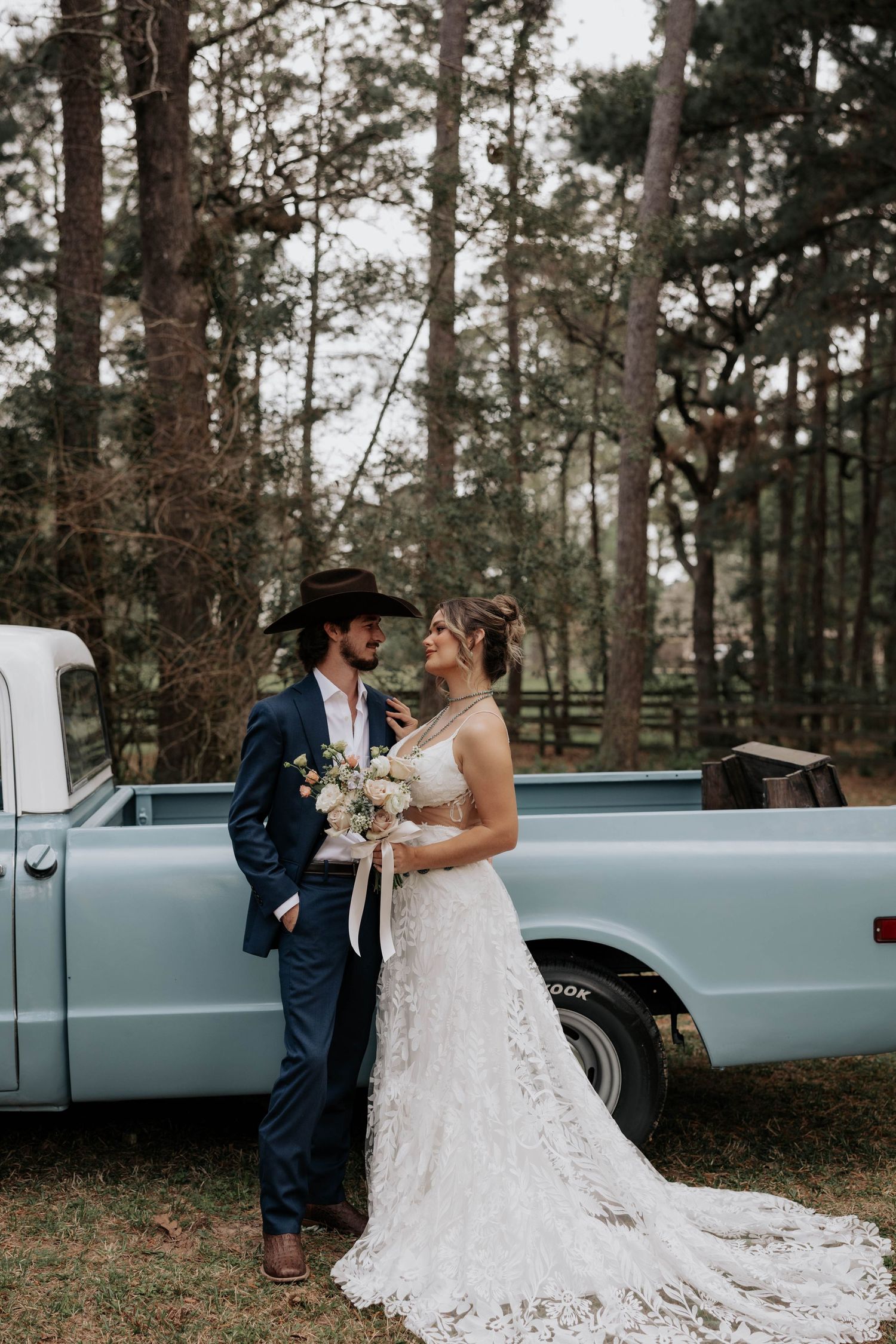 A romantic wedding couple stands by a vintage light blue pickup truck in a pine forest setting.