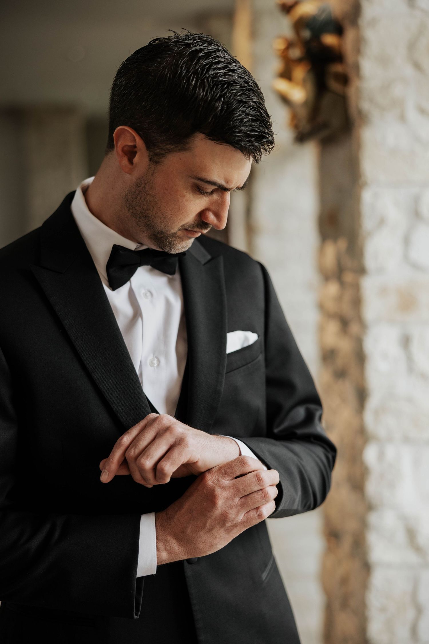 A groom in a classic black tuxedo and bowtie adjusts his cufflinks in an elegant interior setting.