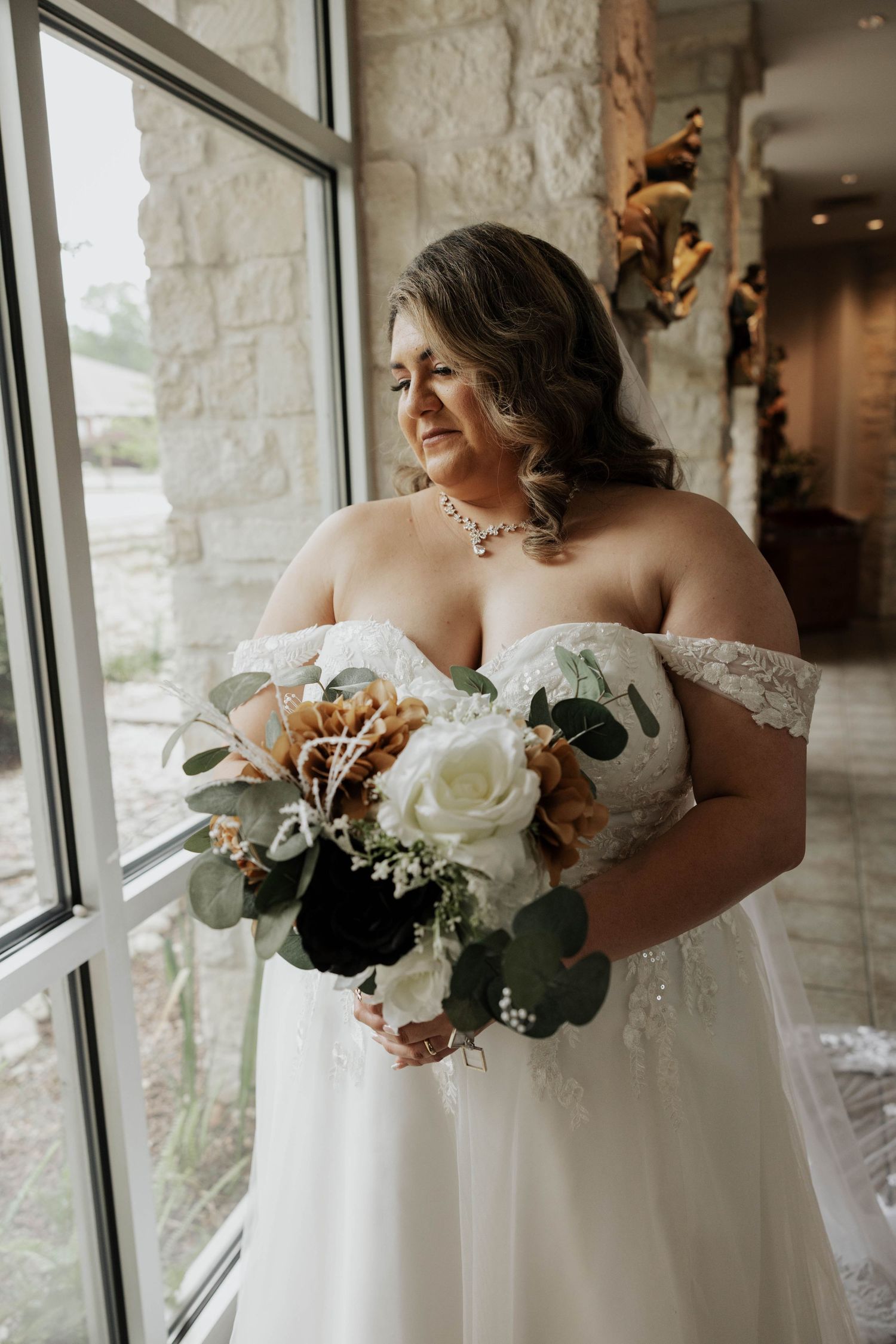 A bride in an off-shoulder lace wedding gown holds a beautiful bouquet of white roses and autumn florals by a window.