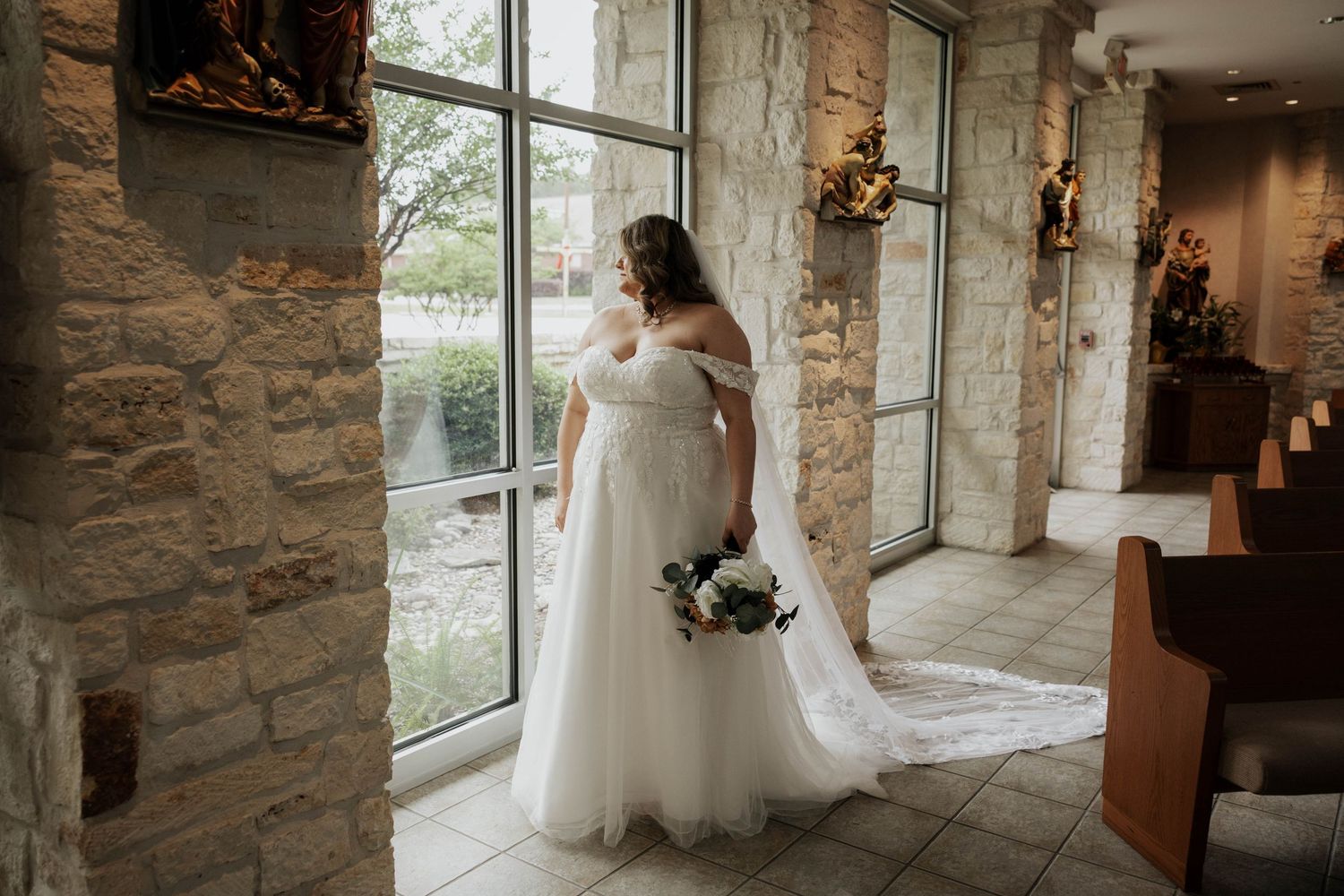 Bride in flowing white gown poses by rustic stone wall interior with natural window lighting.