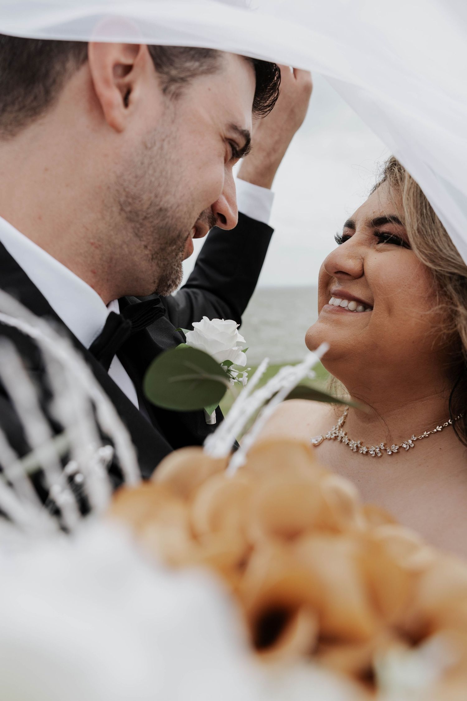 A romantic moment between newlyweds under a veil, with the bride wearing a necklace and a white boutonniere on the groom's lapel.
