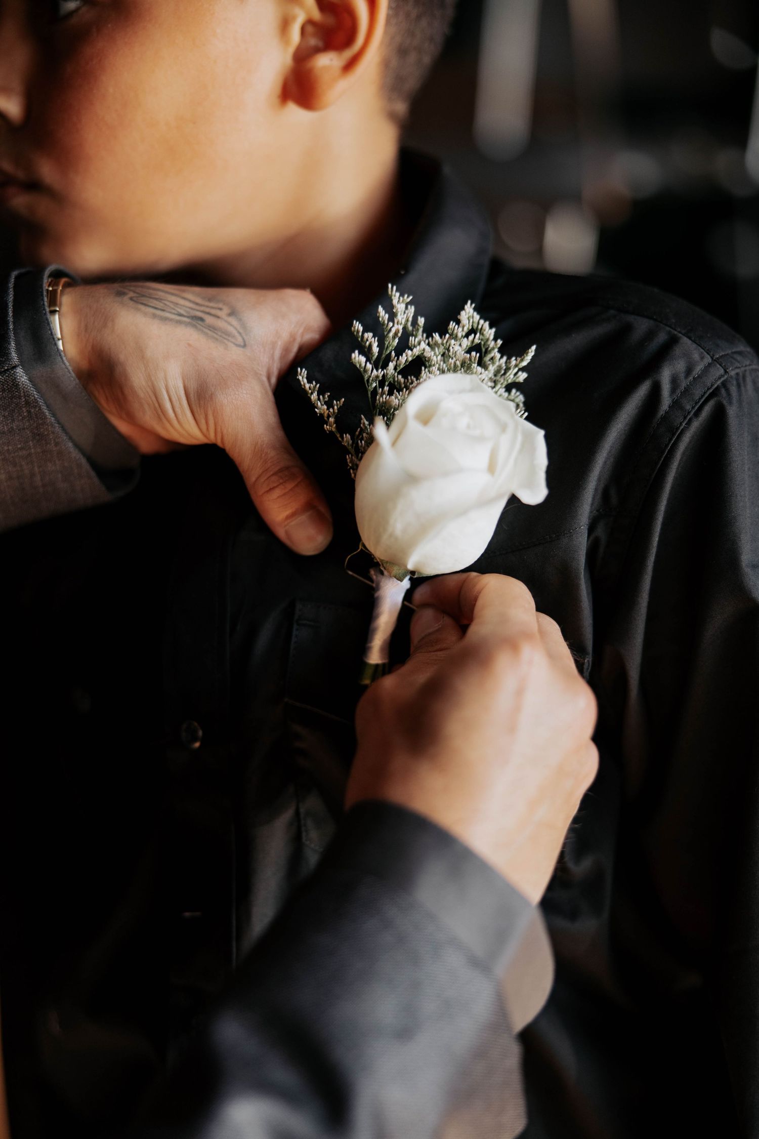 Someone pins a delicate white rose boutonniere onto a dark formal jacket in moody lighting.