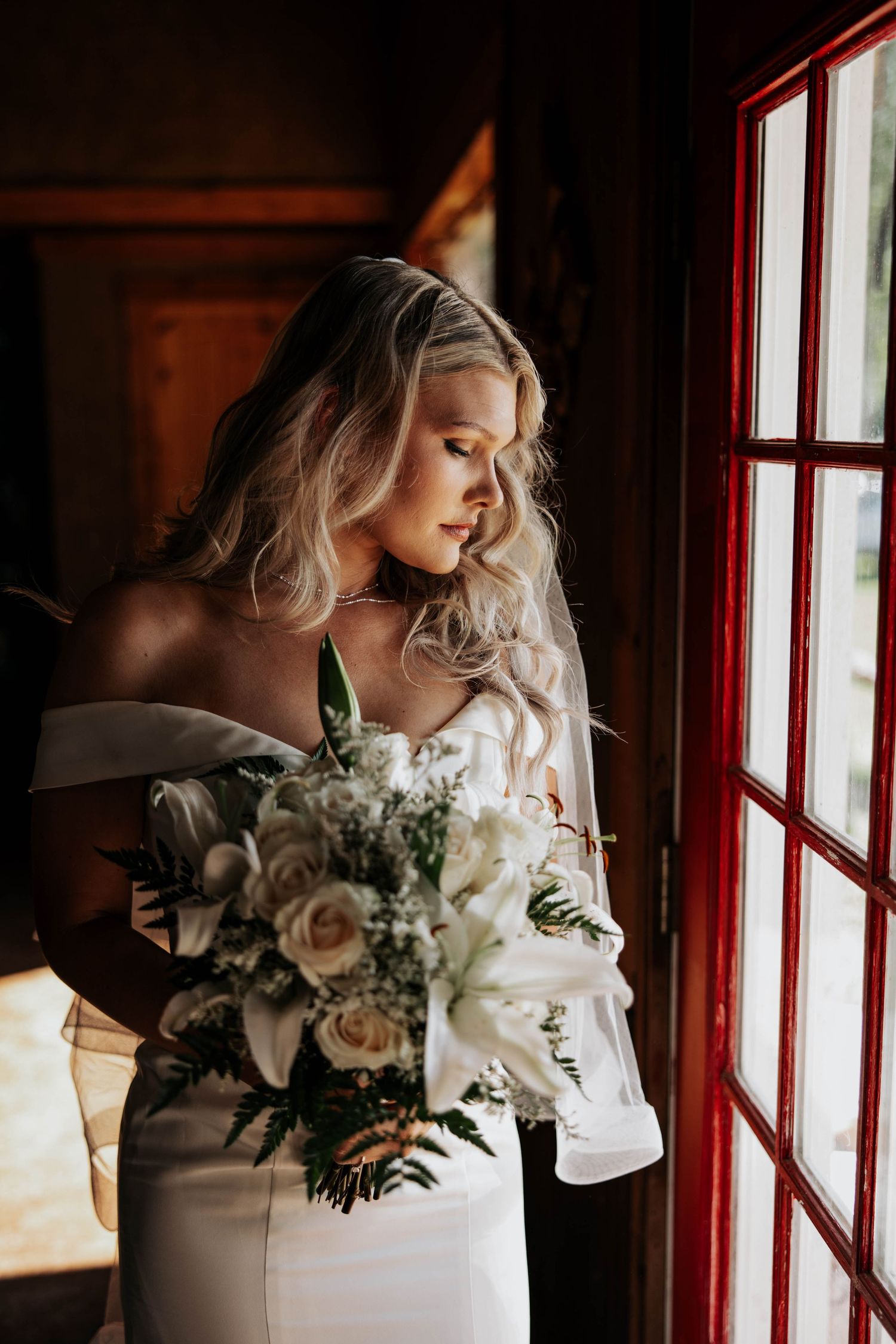 Elegant bride in an off-shoulder white gown holds a romantic bouquet of white roses while standing by a window in warm light.