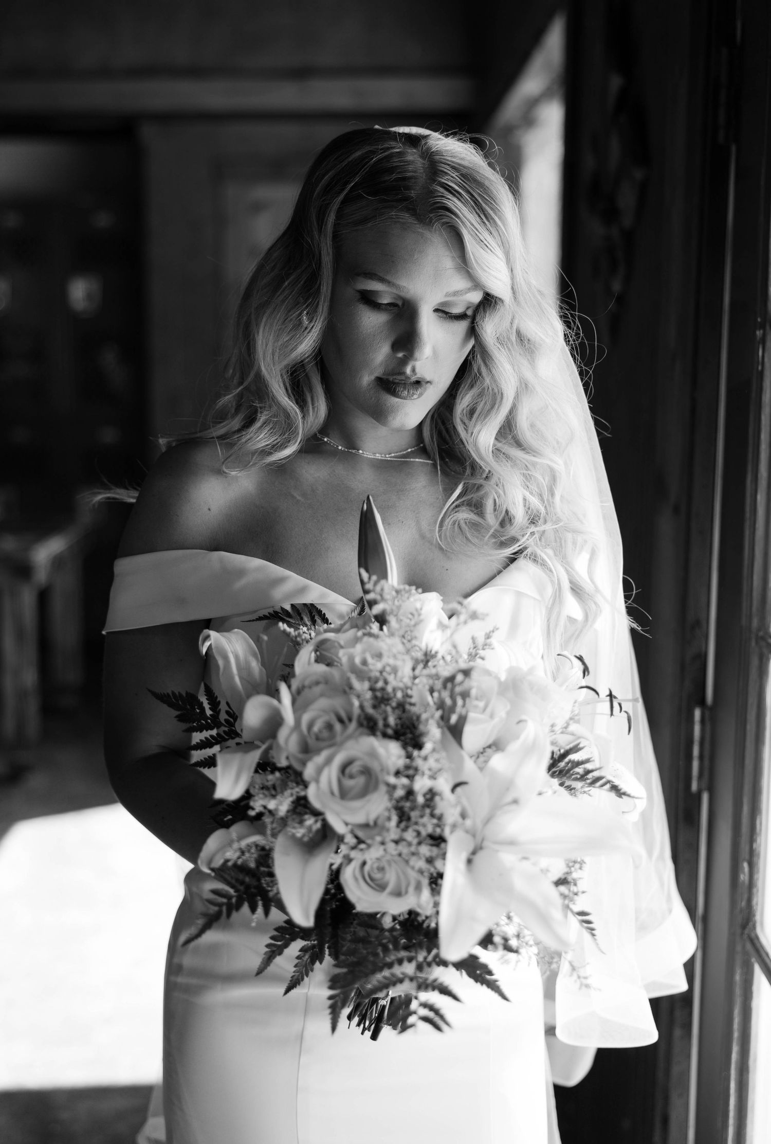 A black and white photograph of a bride in an off-shoulder gown holding an elegant bouquet of roses and lilies.