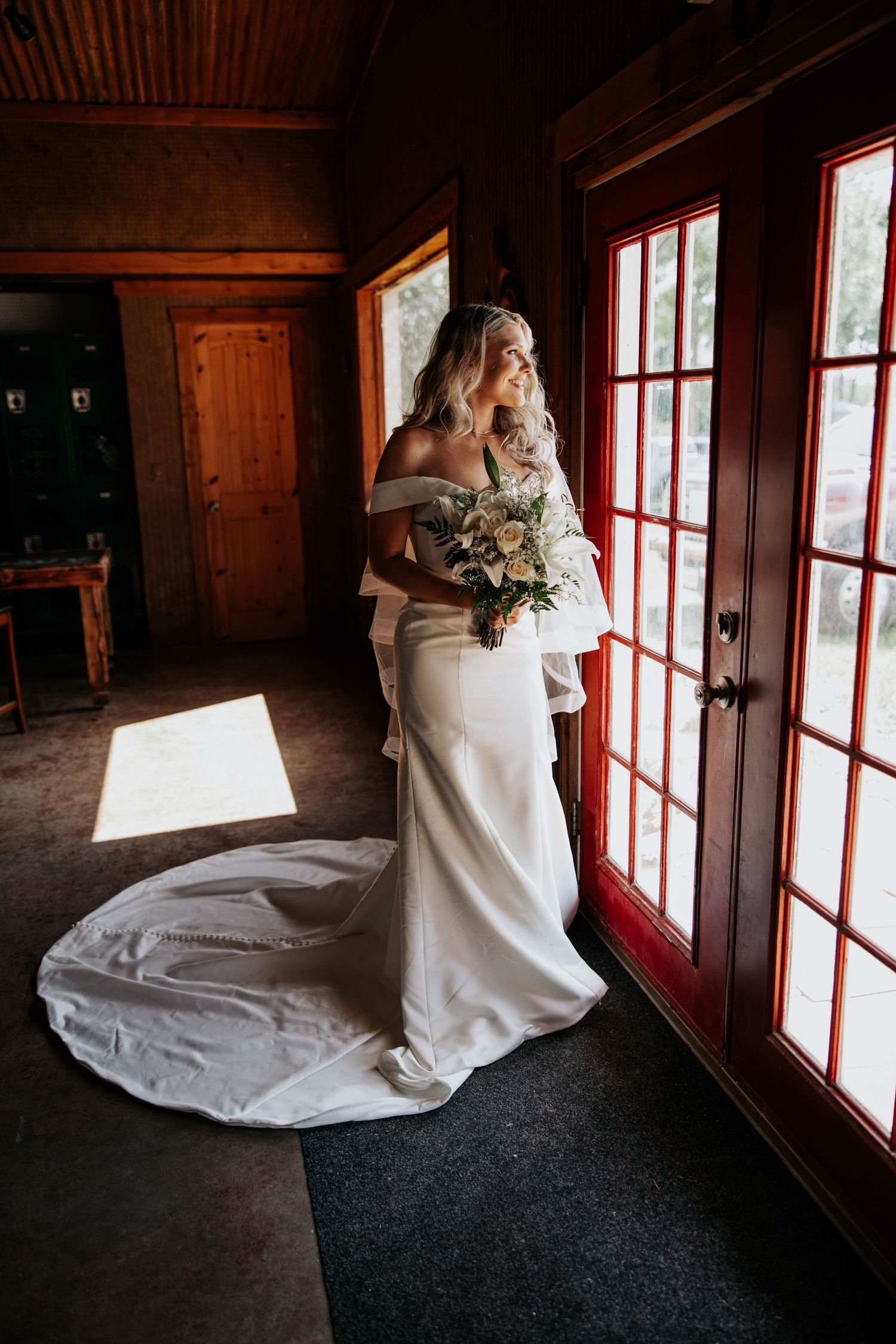 Elegant bride in off-shoulder gown and long train stands by French doors in rustic wooden room with natural light streaming in.