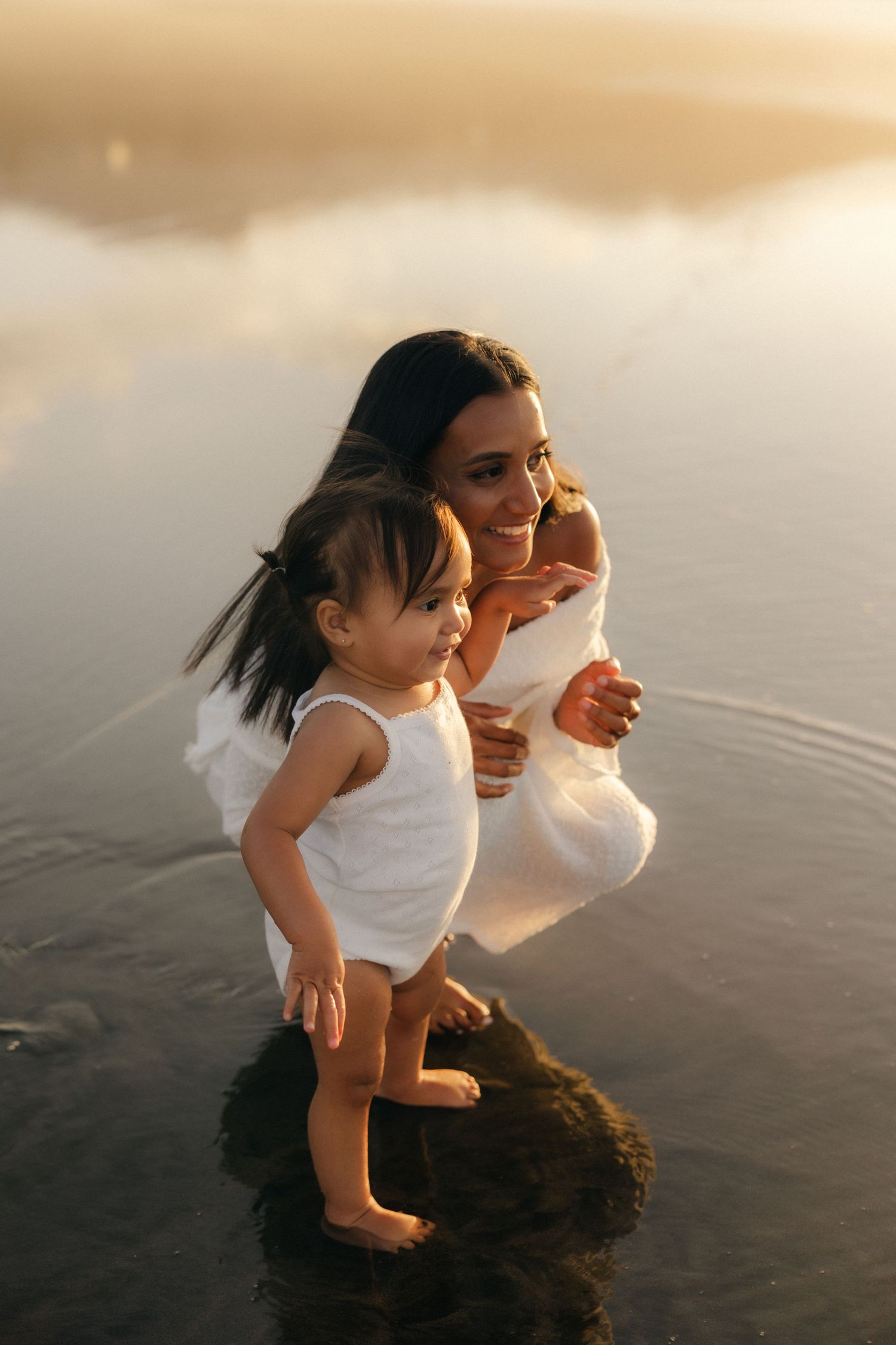 Mother and child sitting on white blanket in grassy field during golden sunset.