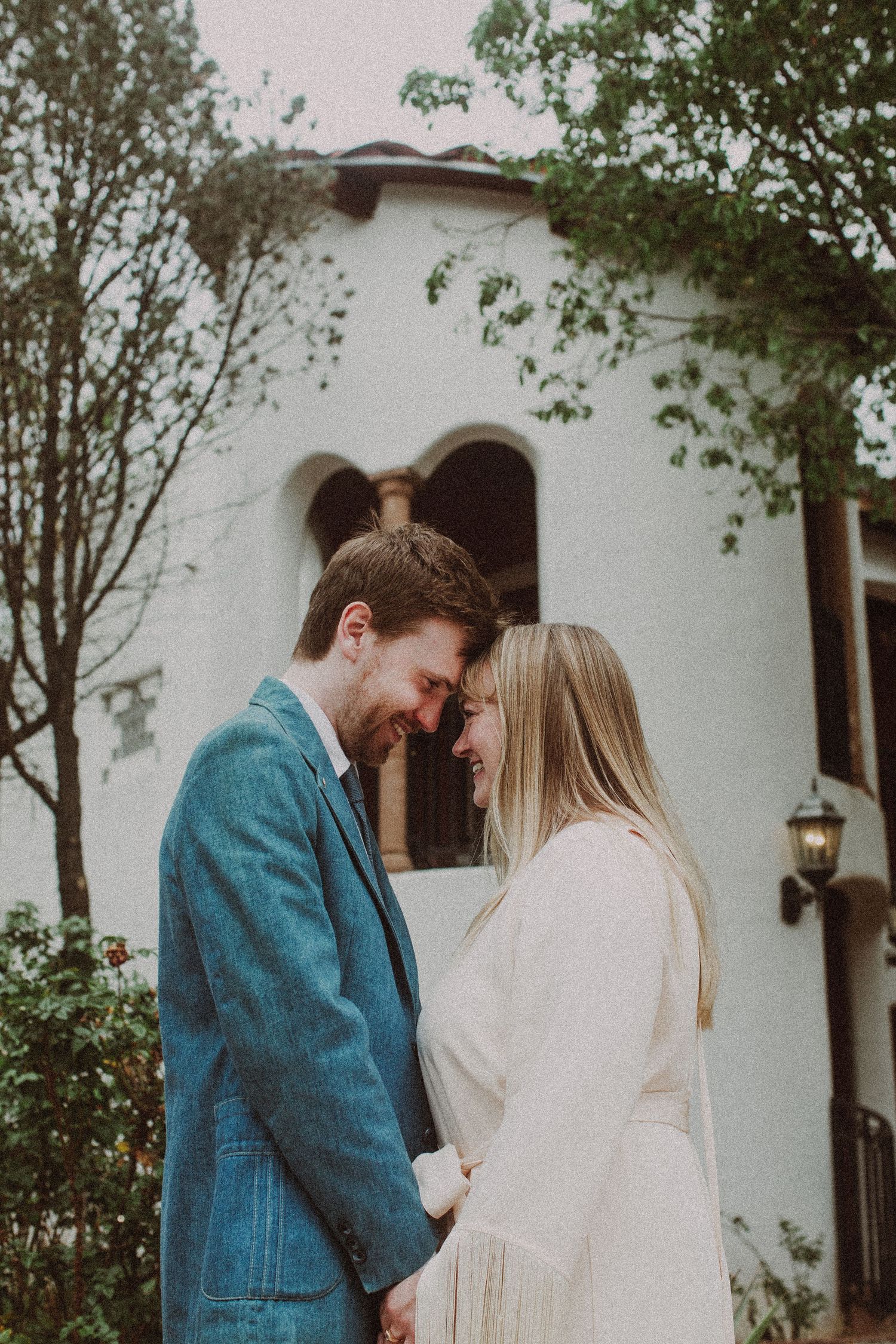 Happy couple in wedding attire share an intimate forehead touch outside white Spanish chapel.