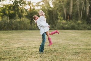 A joyful couple embraces in a grassy field during sunset, with one person lifting the other in a romantic pose.