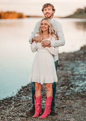 Pink cowboy boots peek out beneath a flowing white dress on a rocky beach at sunset.