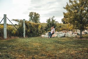 A couple sits together on large rocks in a grassy field during autumn with metal poles and trees in the background.