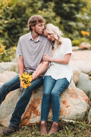 A couple sits together on a large orange-tinted rock outdoors, sharing intimate moments among sunflowers and greenery.