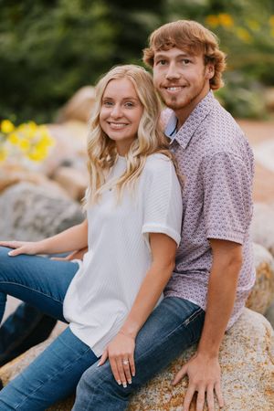 A couple in casual attire sits together on rocky terrain with yellow flowers in the background.