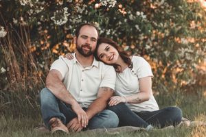 A casual couple in white shirts and jeans sits together on grass during golden hour in a natural outdoor setting.