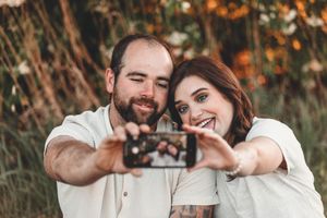 A couple forms a heart shape with their hands while taking a selfie outdoors during autumn.