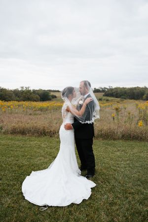 A couple in wedding attire share an intimate dance together in a scenic field with wildflowers at sunset.