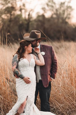 A romantic rustic wedding portrait in tall golden grass with a cowboy hat and lace dress at sunset.