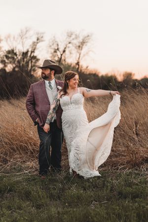 A couple poses in a rustic field at sunset, with the bride's lace dress flowing in the breeze.