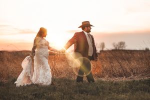 A couple in western attire walks through a golden field during sunset with sunflare in the background.