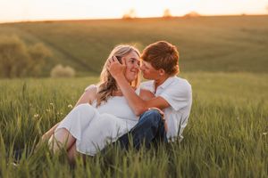 A romantic golden hour portrait of a couple in white clothing sitting together in a tall grass meadow at sunset.