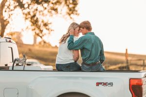 A couple sits together in the bed of a pickup truck watching the sunset in a rustic outdoor setting.