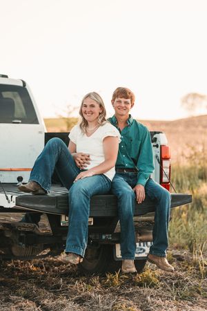 A couple sits together in the back of a pickup truck at sunset in a rural field.