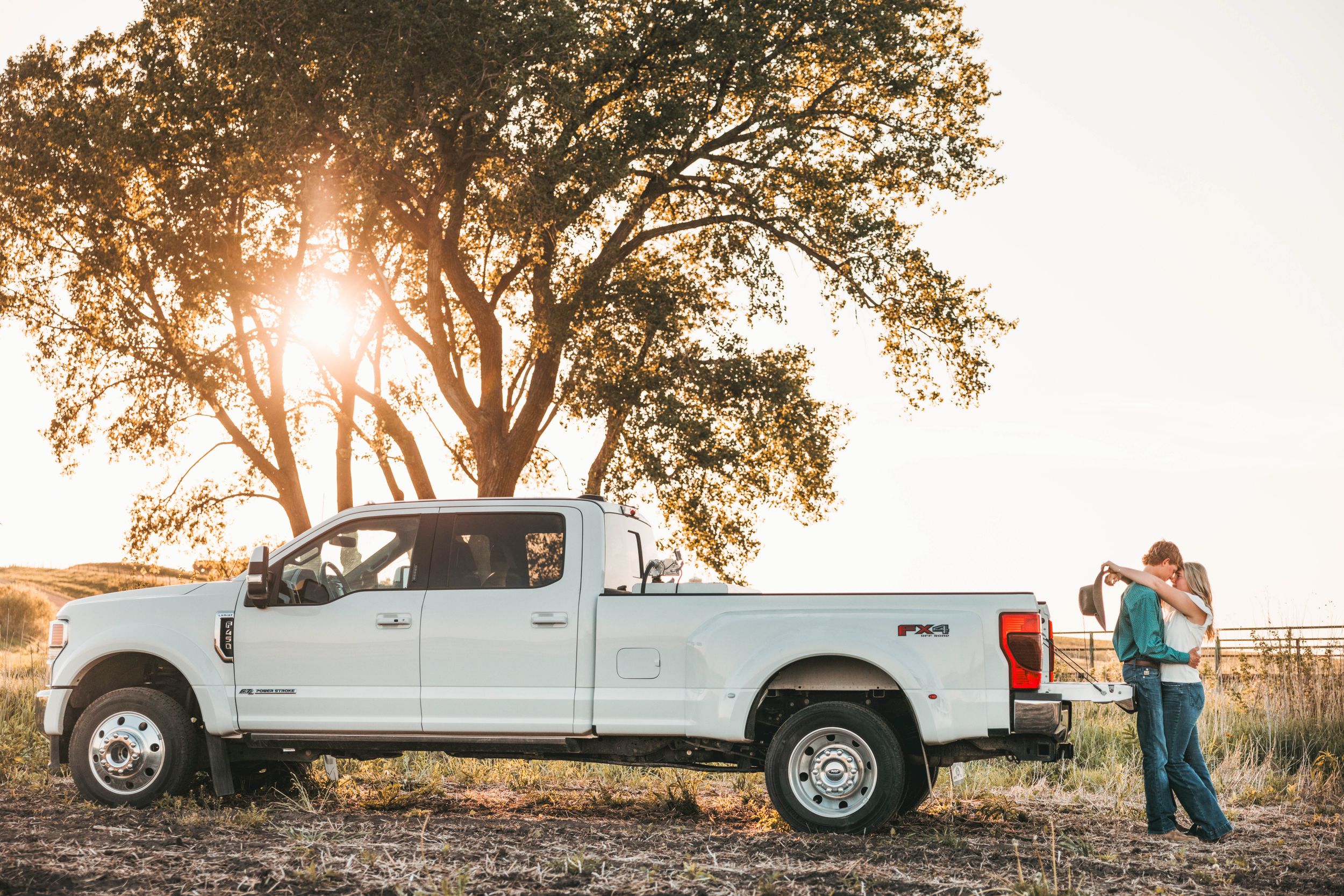 A white Ford Super Duty pickup truck parked near a large tree at sunset with silhouettes in the background.