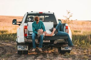 Two people relax on the tailgate of a white pickup truck in an open field during golden hour.