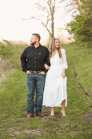 A couple poses outdoors on a green path, man in black shirt and jeans, woman in white lace dress, at golden hour.