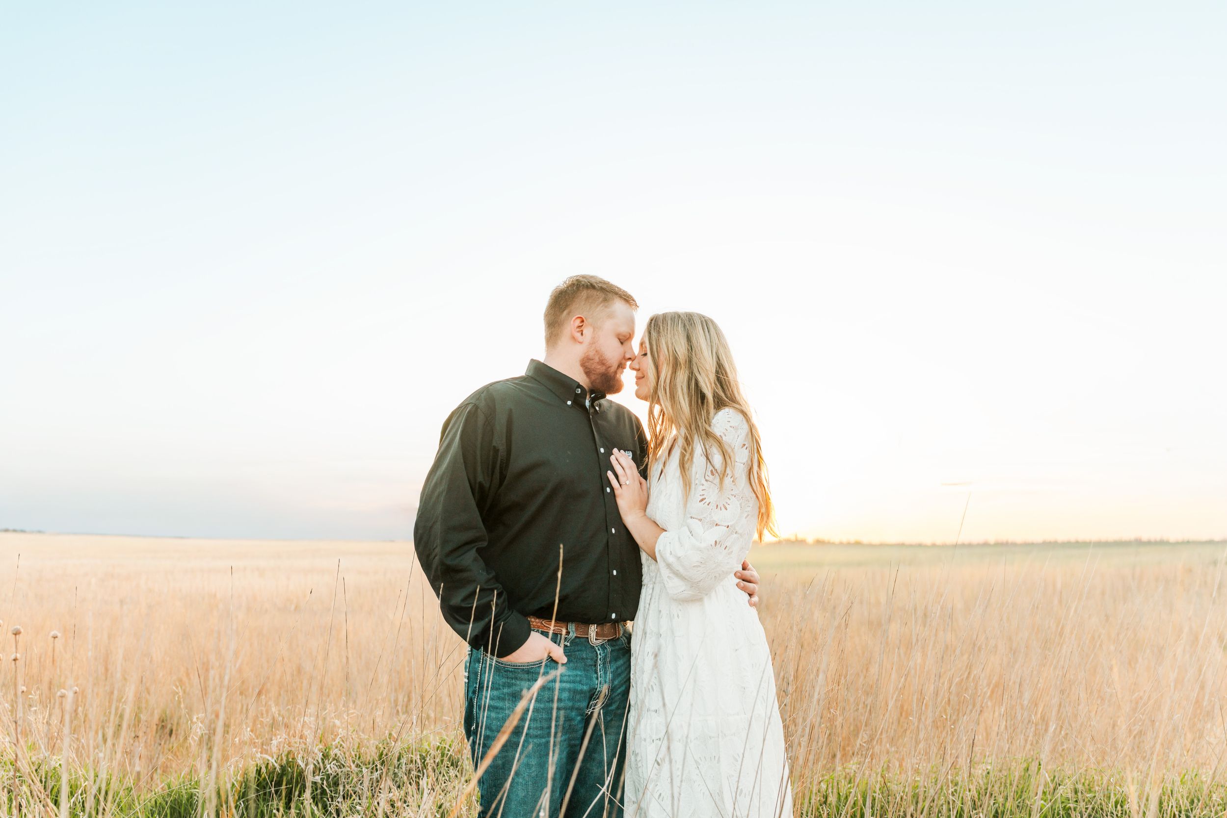 Couple sharing a romantic moment in a golden wheat field at sunset, woman in white lace dress and man in dark jacket.