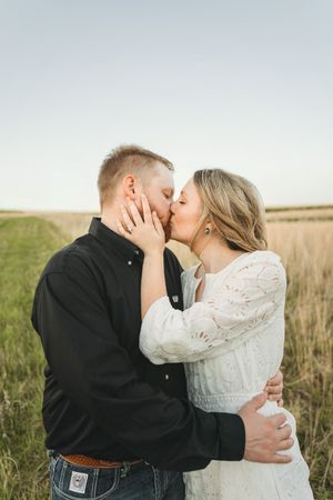 Couple sharing a romantic kiss in a grassy field, man in black shirt and woman in white lace dress embracing outdoors.