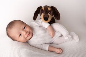 Baby in white knit outfit laying on white surface with plush brown dachshund toy. New Jersey Newborn Photographer
