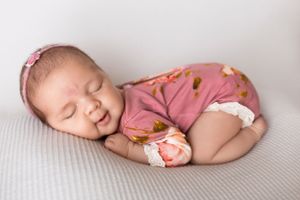 Sleeping newborn baby wearing pink floral outfit curled up on soft gray blanket in New Jersey Studio Photography