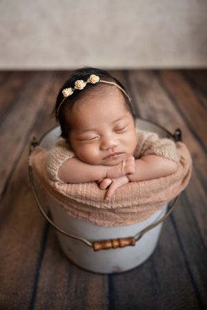 Sleeping newborn baby girl in a metal bucket with a floral headband on a wooden floor photographed by a NJ photographer in Morris County NJ