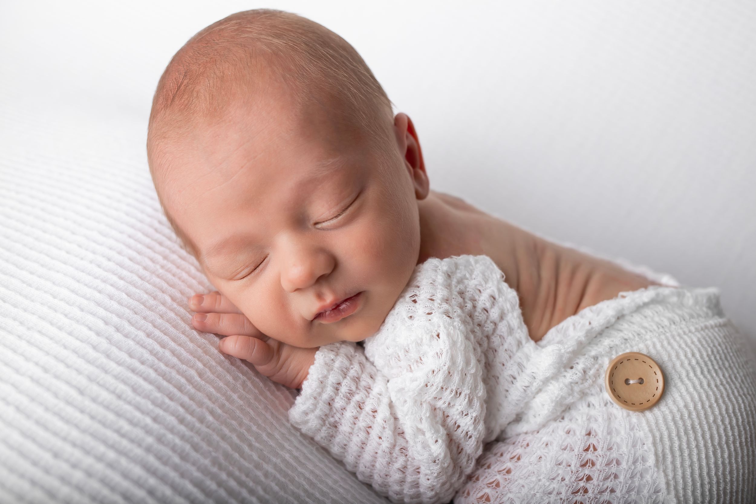 Sleeping newborn wearing white knit sweater on textured white surface.