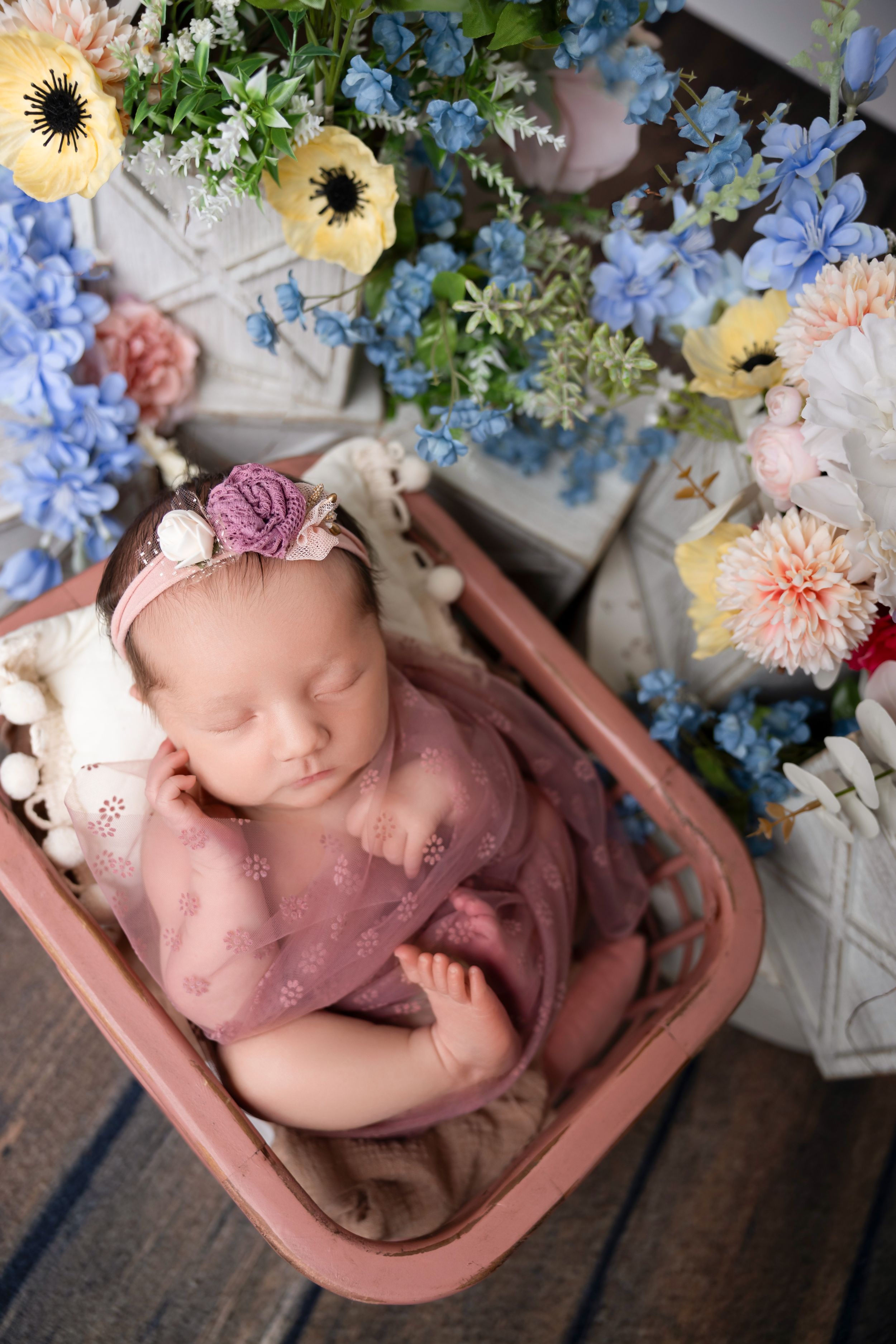 newborn wrapped in purple swaddle with floral headband in pink basket surrounded by colorful spring flowers. NJ newborn photographer