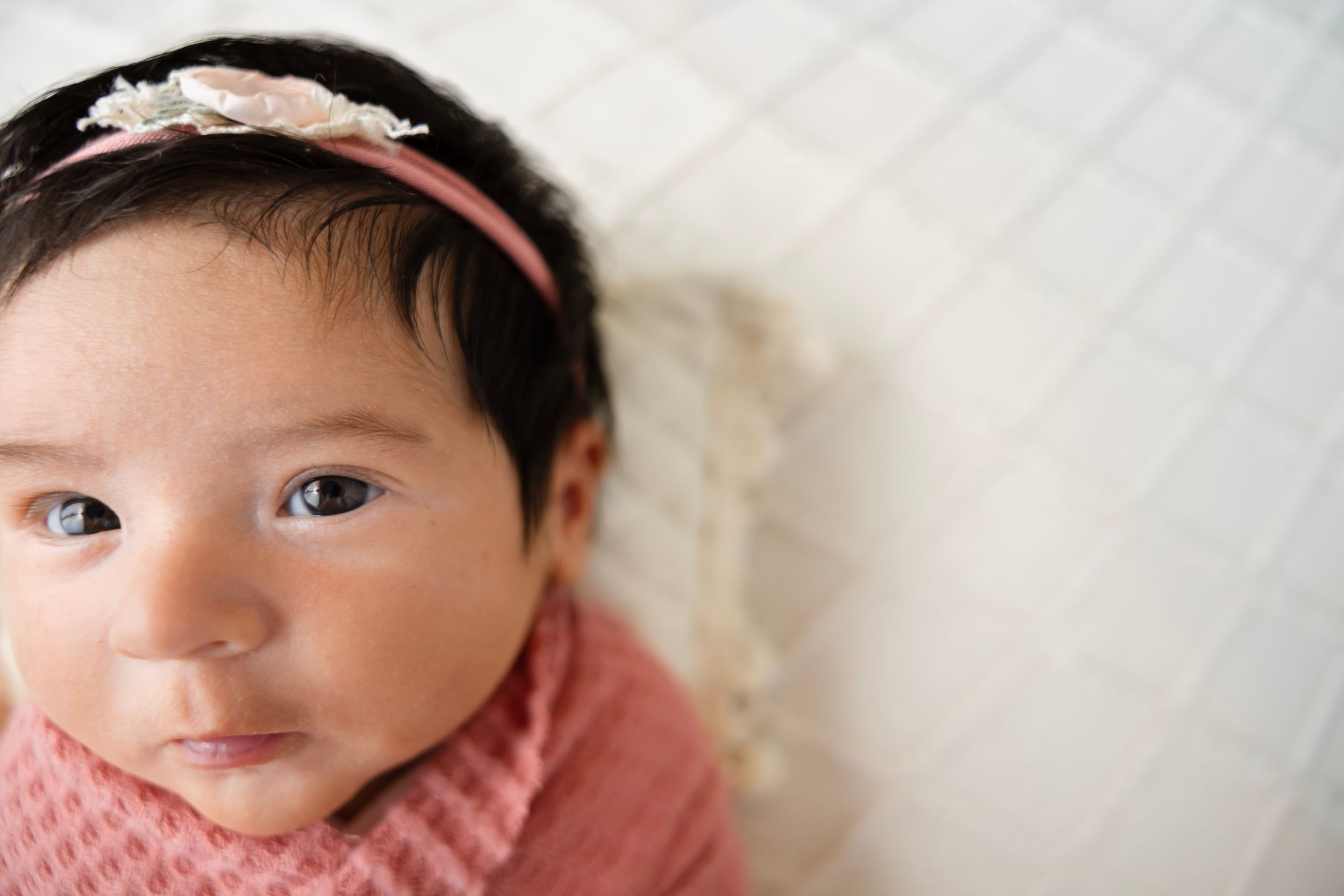 Newborn baby girl wearing a pink floral headband wrapped in a pink knit blanket looking up at the camera.