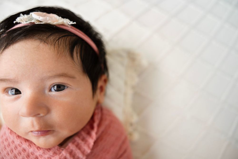 Newborn baby girl wearing a pink floral headband wrapped in a pink knit blanket looking up at the camera.