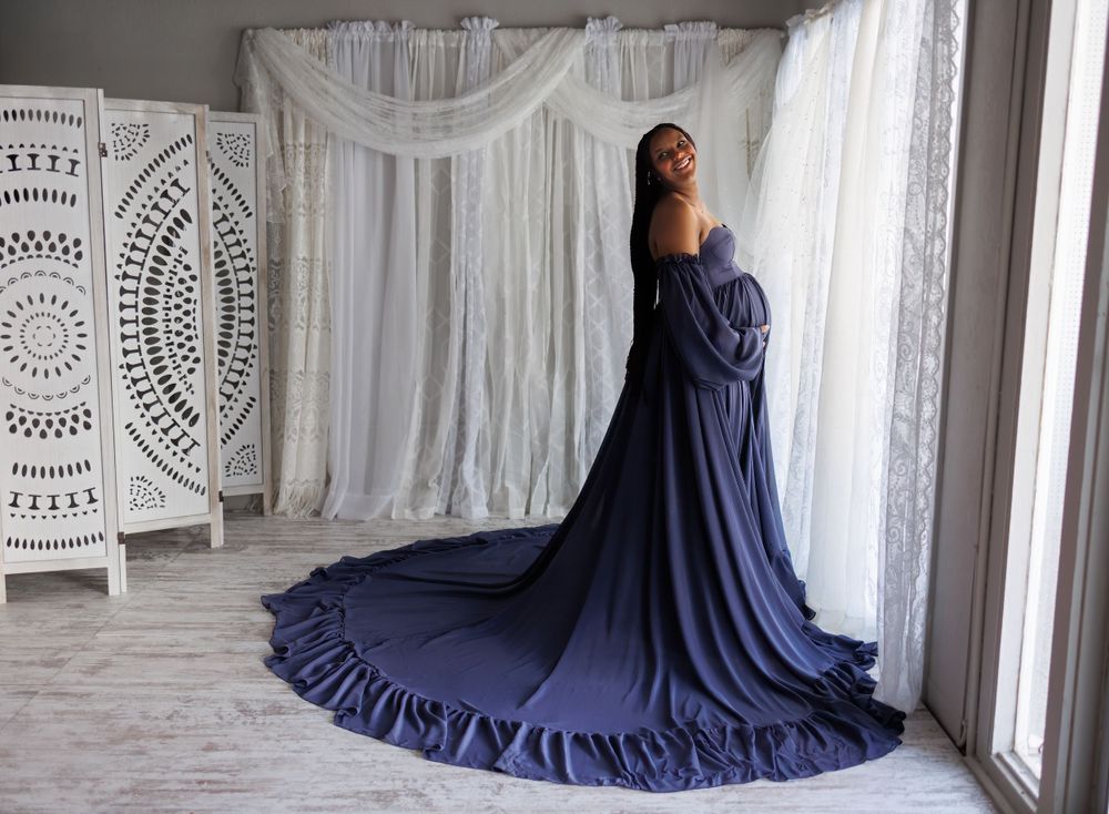 Woman in flowing navy blue gown posing against white curtained backdrop.
