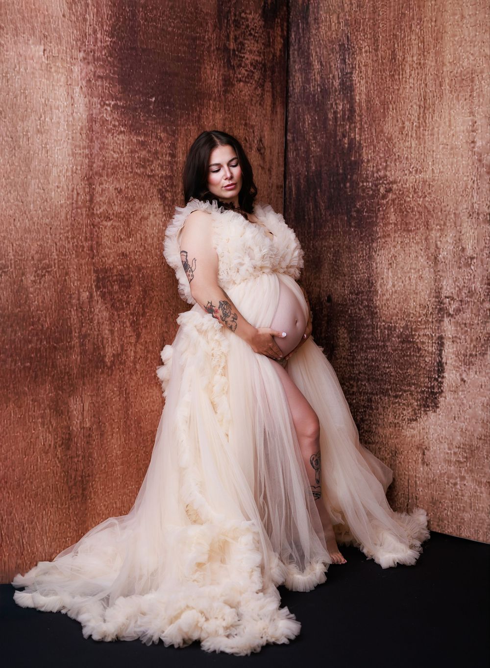 A pregnant woman in a flowing cream tulle gown poses against a rustic textured brown wall in an artistic maternity photo.