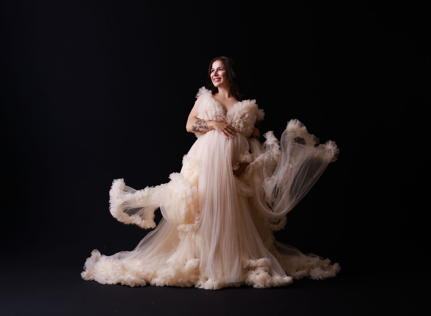 A flowing blush-colored gown with ethereal fabric billowing dramatically against a dark background in a maternity photoshoot.
