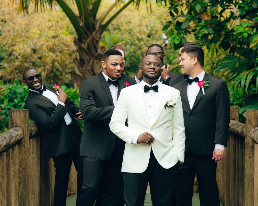 Wedding party wearing black tuxedos and white jacket pose together in lush garden setting.