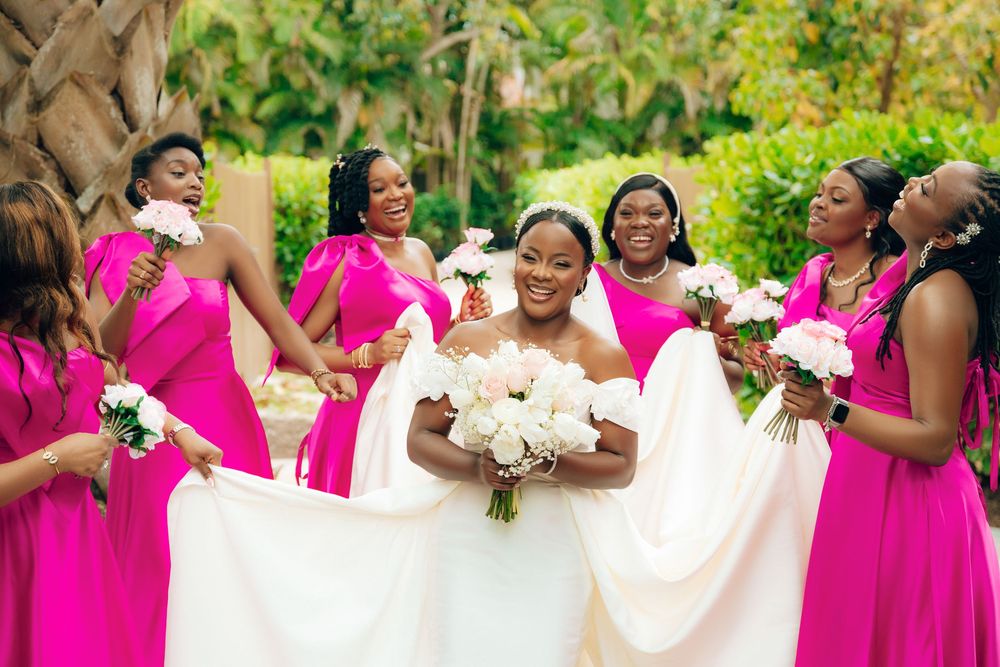 A wedding party wearing bright pink bridesmaid dresses stands outdoors with bouquets as they celebrate together in a garden.